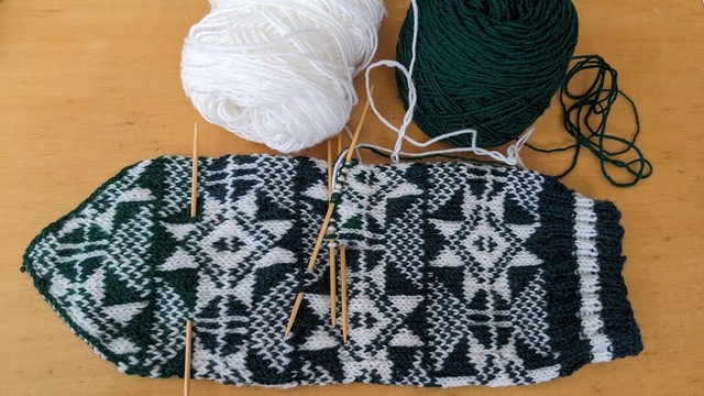 Image of a tubular knitted object made of dark green and white yarn. It is knitted with similar but different colored yarns from the middle.