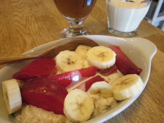 Close up on a dish of porridge with a splash of soy milk, and topped with slices of banana and plum.  A glass of iced tea and cold soy milk in the background