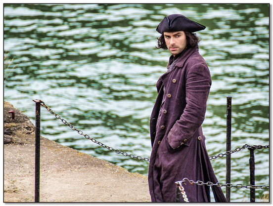 The Actor, Aidan Turner in costume as Ross Poldark, walking across a dock, with the sea in the background.