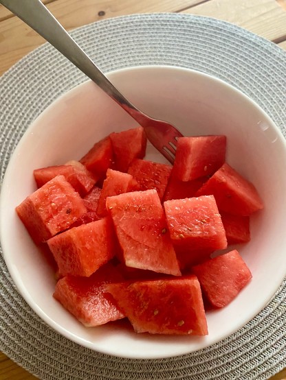 A white bowl of cut up chunks of red seedless watermelon and a fork on a circular ridged placemat 