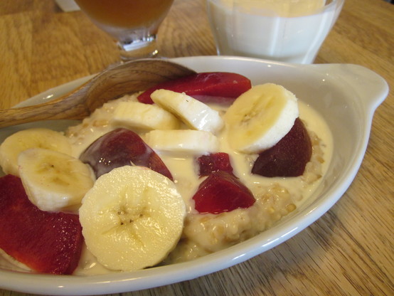 A close up shot on a dish of porridge with a splash of soy milk, with topping of sliced banana and plum.  A glass of iced tea and another of cold soy milk in background