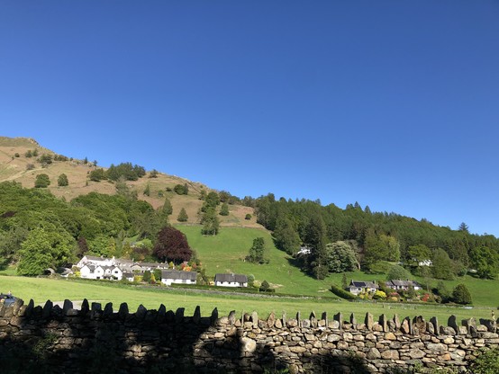 イングランドのカンブリア地方グラスミアの田園風景。石垣で仕切られた野原、白い壁のコテージ。青空と丘の緑のコントラストが美しい。
A scenic countryside view in Grasmere, Cumbria region, England, with a stone wall in the foreground, green fields, white cottages, and a forested hillside under a clear blue sky.