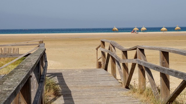 Ein Holzsteg mit Geländern auf beiden Seiten, der hinunter zu enem breiten Sandstrand führt. Der Strand ist fast menschenleer. Nur zwei Personen liegen in einiger Entfernung unter einem von insgesamt fünf Sonnenschirmen aus Stroh. Dahinter erscheint das Meer in einem schmalen, blauen Streifen. Darüber blauer Himmel.