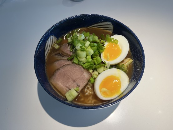 A blue and white bowl with a partially visible fish decoration. In it can be seen a brown broth with green onion, slices of pork, a soft-boiled egg sliced in halves and some ramen noodles below.