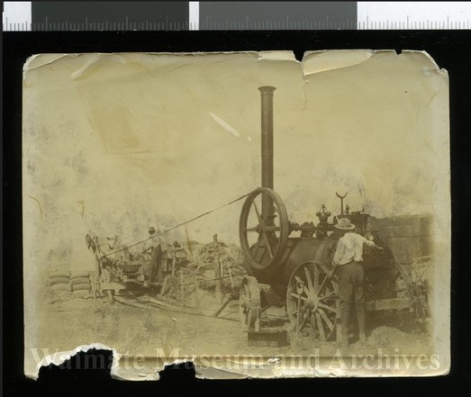 Black-and-white photo: Stationary steam engine and chaff cutter, Hakataramea. 1920s? Description: Shows a man standing beside a stationary traction engine that is connected by a long belt to a chaff cutter at which another man is working. Both men are wearing long trousers, long-sleeved, light-coloured shirts and hats. Citation: Waimate Museum & Archives, Catalogue No. P8532.081. https://waimate.pastperfectonline.com/Photo/B525303A-3B78-469D-9786-384742462632