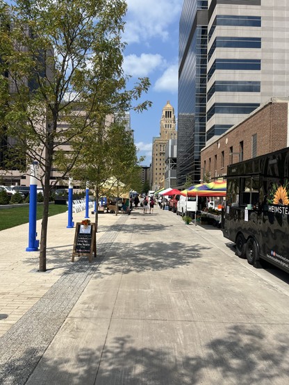 View of a street. The street is open to pedestrians. On either side, tent pavillions. Tall downtown buildings can be seen farther down. A blue sky dominates with a few puffy white clouds. Trees line the street, giving a little shade. Green grass off to one side.