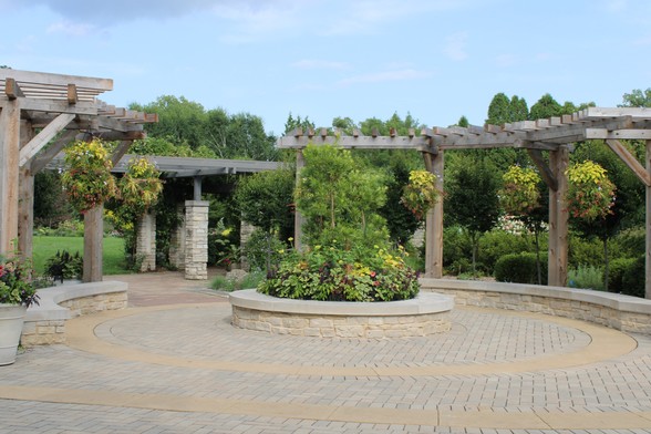 A circular large stone wall edged raised flower bed filled with flowers and a tree with pavers laid in a circular pattern between the flower bed and two semi circular stone benches supporting wooden beamed arbors with hanging plant displays to the left and right of the central circular bed with a walkway between leading to an arbored walkway with stone pillars in front of a large lawn area. There are trees and an assortment of flora in the background under a blue sky with wispy clouds.