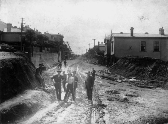 Black-and-white photo: Widening Wallace Street, Mt Cook, Wellington, for trams. 1925. Photographer unidentified. Description: Wallace Street, Mt Cook, Wellington, being widened for trams in 1925. A group of construction workers are in the foreground. Prior to this time, the tramway went only as far as Howard Street. No 149 is on the right, marked with a cross. Both 149 and the house next door were built by Ovid Dempsey. Citation: Original photographic prints and postcards from file print collection, Box 7. Ref: PAColl-6001-13. Alexander Turnbull Library, Wellington, New Zealand. https://natlib.govt.nz/records/22407002