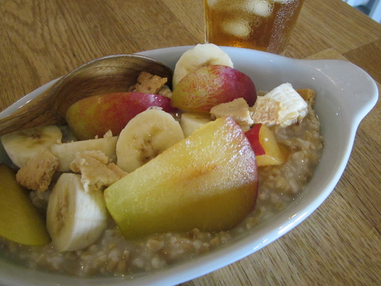 Close up on a dish of porridge with toppings of banana, plum and crumbled Italian honey biscuit.  A glass of iced tea in the background.