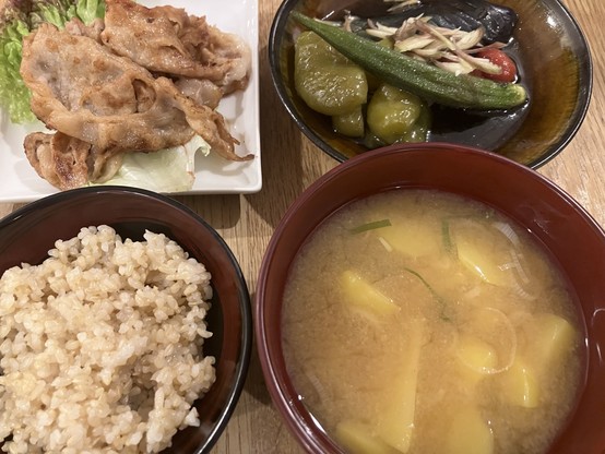 A meal consisting of four dishes: grilled meat on a plate with lettuce, a bowl of mixed vegetables including okra and eggplant, a bowl of brown rice, and a bowl of miso soup with potatoes.