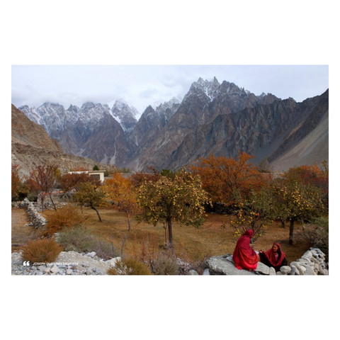 Photo of a small fruiting apple orchard backgrounded by the icy and towering peaks of Passu Cones in Gilgit-Baltistan, Pakistan. Two veiled local women can be seen in the corner in their bright red traditional dress.