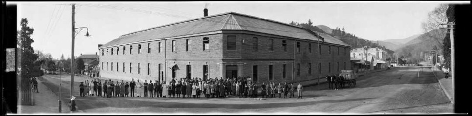 Black-and-white photo: Staff of Griffin’s Biscuit Factory outside the factory building in Nelson. 1920s. Photographer, Robert Percy Moore. Description: Staff of Griffin & Sons Biscuit factory standing outside the factory building in Nelson. Dray drawn by two horses in the street at left. Photograph taken between 1923 and 1928. Citation: Moore, Robert Percy, 1881-1948: Panoramic photographs of New Zealand. Ref: Pan-0154-F. Alexander Turnbull Library, Wellington, New Zealand. https://natlib.govt.nz/records/30660014