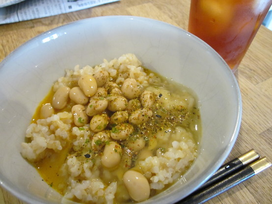 An earthenware bowl of brown rice topped with a mixture of raw egg and marinated soybeans.  A glass of iced tea and a folded Japanese newspaper in the background