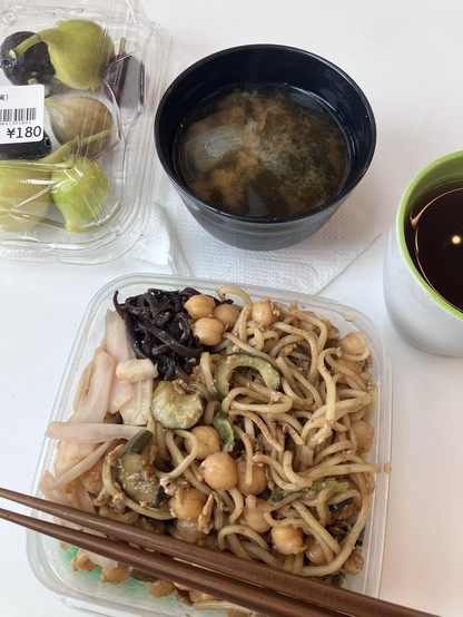A meal consisting of a plastic container with noodles, vegetables, and chickpeas, a cup of miso soup, a cup of tea or coffee, and a packaged container of figs.