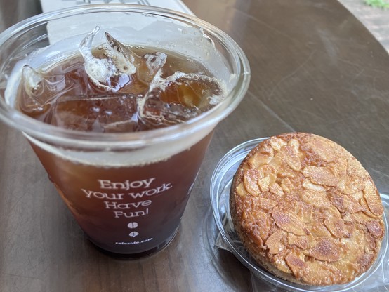 A plastic cup of iced coffee next to a round almond pastry on a dark wooden table. The cup has the text 