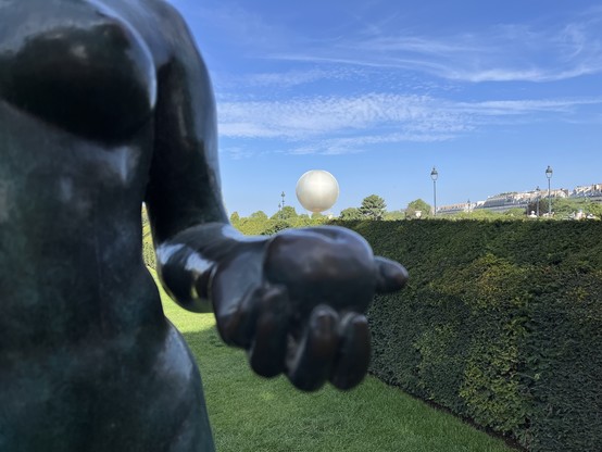 Dans les jardins du Louvre, au premier plan une statue d'une femme tenant une pomme et juste au dessus la vasque dorée Olympique se détache en arrière plan.