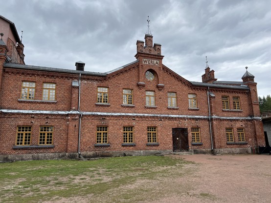 Historic brick building with yellow-framed windows under a cloudy sky. The building has turrets and a central clock. 