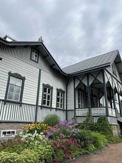 A wooden house with a decorative gabled roof, surrounded by a colorful garden in bloom under a cloudy sky.