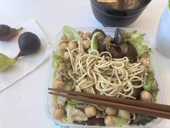 A close-up of a meal with a plastic container of noodles, chickpeas, and vegetables, paired with wooden chopsticks. In the background are two figs, a bowl of soup, and a drink.