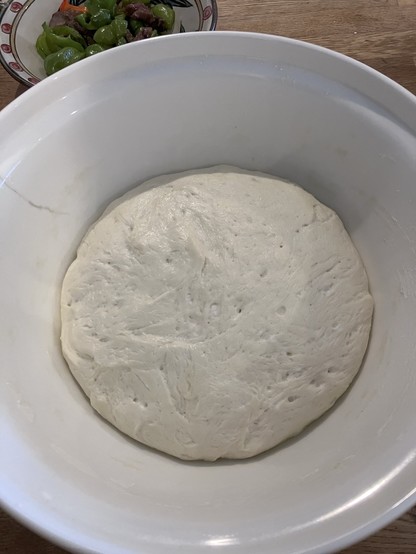 A bowl of risen dough on a wooden surface, with a plate of sliced green peppers and other vegetables in the background.