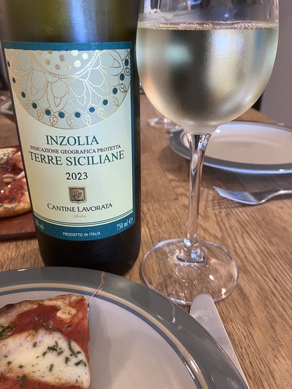 Bottle of Inzolia Terre Siciliane 2023 wine with a glass of the same wine and a slice of pizza on a plate in the foreground.