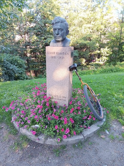 A bust of Oskar Braaten on a plinth surrounded by flowers. There is a unicycle leaning on the plinth.