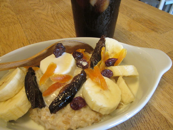 Close up on a dish of porridge with splash of soy milk, topped with sliced dates, dried apricots, banana and dried cranberries.  A glass of iced coffee in background.