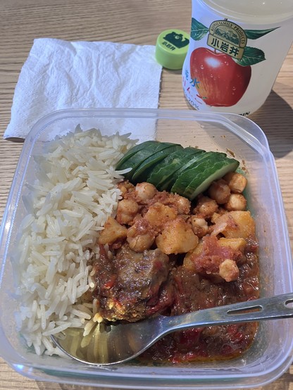 A meal container with rice, chickpeas, potatoes, sliced cucumbers, and a stew, along with a spoon. A bottle of apple-flavored drink and a napkin are placed next to it.