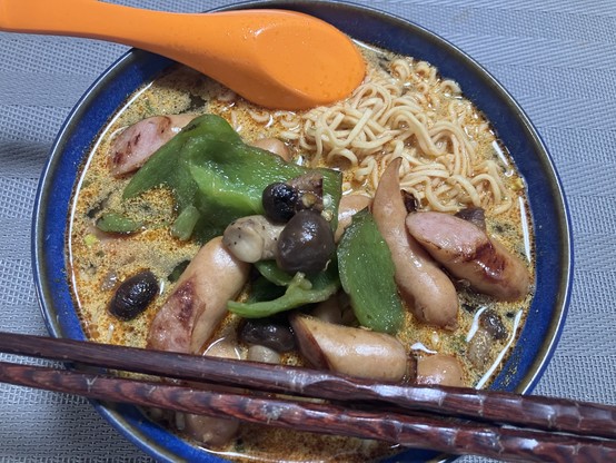 A blue bowl containing noodle soup with chunks of sausages, green bell peppers, mushrooms, and an orange spoon. Two wooden chopsticks are placed beside the bowl.