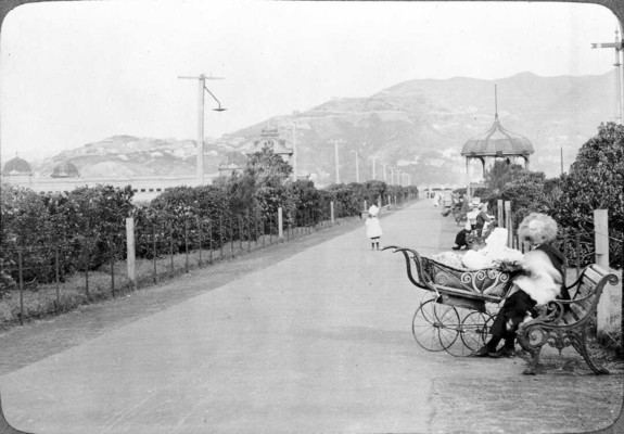 Black-and-white photo: Thorndon Esplanade, Wellington. ca. 1910. Photographer unidentified. Description: Looking down the esplanade, which is edged with shrubs. A young girl stands in the middle of the path mid-distance; closer and to the right someone sits on a seat next to a pram and probably holding a baby (their image is blurred). More seats with people sitting on them are visible further down the right side of the esplanade. The public baths with their distinctive domes are to the left in the middle distance. In the far distance is Mount Victoria. Citation: Ref: 1/2-106870-F. Alexander Turnbull Library, Wellington, New Zealand. https://natlib.govt.nz/records/23203459