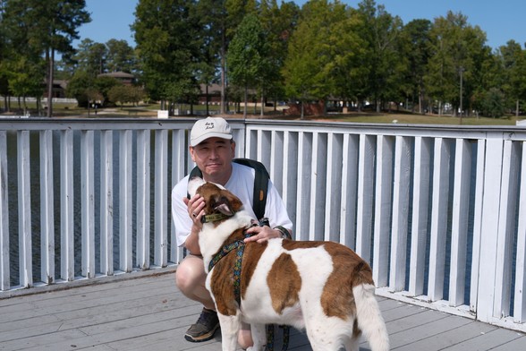 A man with a white cap crouching on a deck over a pond, holding his white and brown dog. White fences, pond, and trees in the background