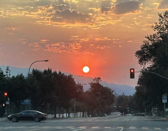 Sun rising above the San Gabriel mountains with clouds lit above. Trees, a road intersection, a red light and a car crossing intersection from right to left in the foreground 