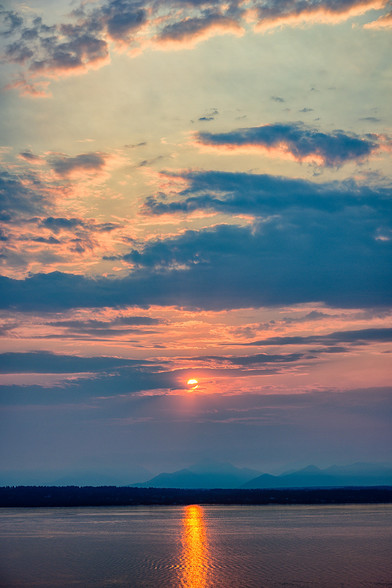 Color photo.  The setting sun, partially obscured by clouds, just above a range of mountains.  At the bottom of the frame is a body of water, upon which is a lane of reflected sunlight.  Above the setting sun, the sky is full of dark clouds, except for a patch of pale blue sky.