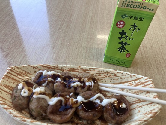 The image shows a plate of bite-sized takoyaki drizzled with sauce and mayonnaise, accompanied by a carton of unsweetened green tea. Two chopsticks are placed on the side of the plate. The setting features a wooden table surface.