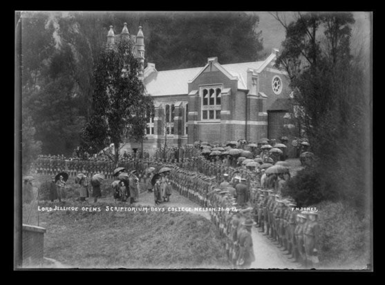 Black-and-white photo: Lord Jellicoe opens Scriptorium–Boys College. Nelson. 26.9.24. Photographer, F. N. Jones. Description: View of a large group of boys in cadet uniforms lining the pathway in front of the Nelson College Scriptorium. Other people gather around, many with umbrellas. Citation: Nelson Provincial Museum, F N Jones Collection: 321266. https://collection.nelsonmuseum.co.nz/objects/P22300/lord-jellicoe-opens-scriptorium-boys-college-nelson-26924