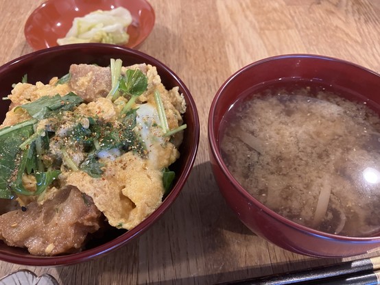 A bowl of rice topped with egg, pieces of fried bread, and greens, garnished with spices, accompanied by a small dish of pickled vegetables and a bowl of clear soup with shiitake.