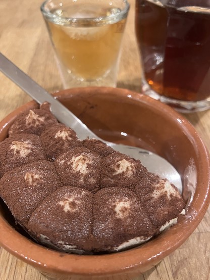 A serving of tiramisu in a clay dish, accompanied by a spoon. Two glasses are visible in the background: one contains a light-colored liquid and the other a dark beverage.