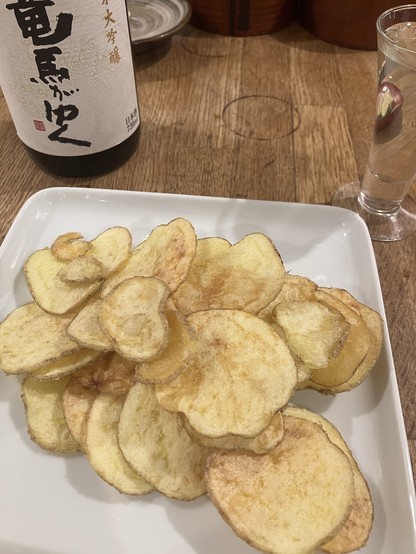 A plate of golden-brown potato chips is served alongside a bottle of sake and a glass. The background features wooden textures and a traditional bowl.