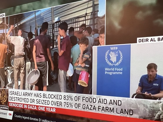 An image of a news broadcast featuring a crowded scene where individuals are waiting for food aid, with some holding cooking utensils. The screen also shows a graphic detailing the impact of the Israeli army's actions, stating that 83% of food aid has been