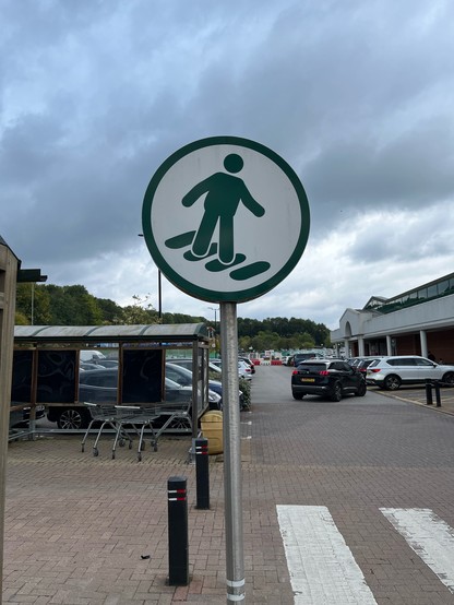 Sign next to a pedestrian crossing in a supermarket car park. It shows a stylised person apparently facing across the narrow width of the crossing, the stripes of which are shown as elongated ovals. The effect is to suggest that the figure is wearing enormous shoes.