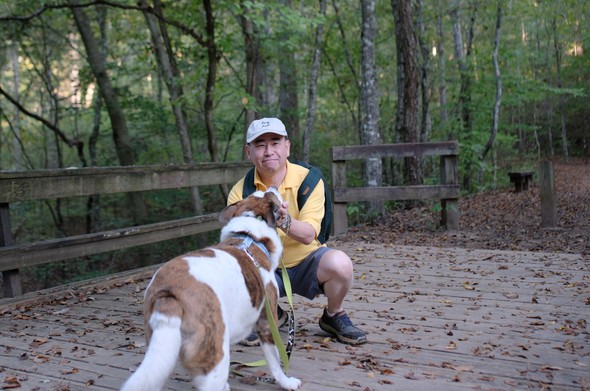 A man with a white cap and a yellow shirt crouching on a wooden bridge, holding his white and brown dog's face