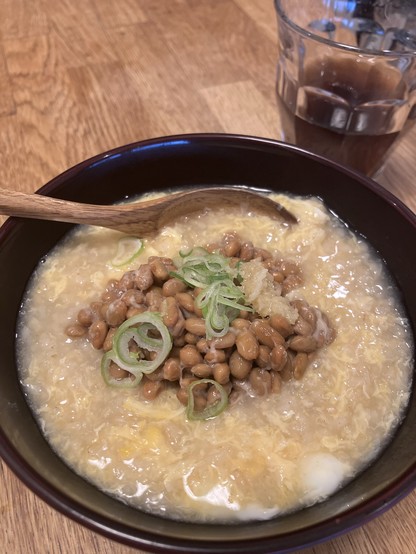 A bowl of warm, creamy rice topped with natto (fermented soybeans), sliced green onions, and a dollop of ginger scallion sauce. A wooden spoon rests on the edge of the bowl, which is set on a wooden surface. In the