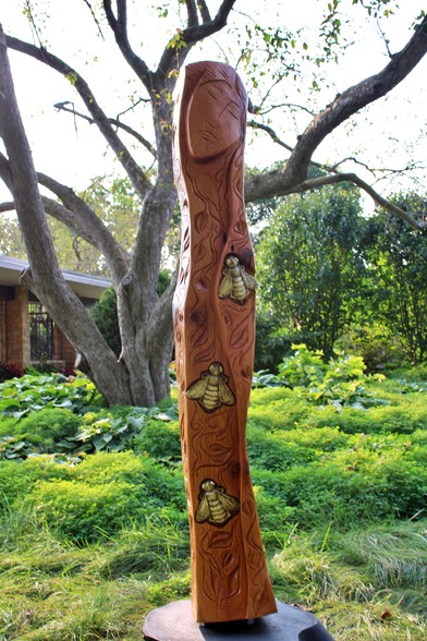 Tall red wood sculpture with the face of a woman at the top with golden bees below with curved lines in the shape and integrated into the carving around the bees. There are plants and trees and a wall with open air windows and a roof in the background. 