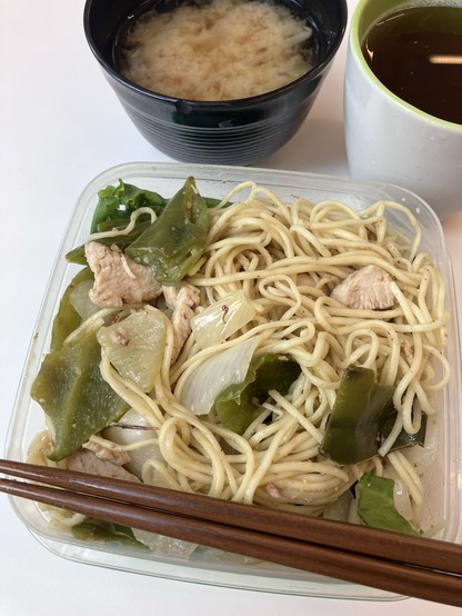 A plastic container filled with stir-fried noodles, pieces of chicken, and green bell peppers, accompanied by a bowl of miso soup and a cup of green tea. Brown chopsticks rest on the side.