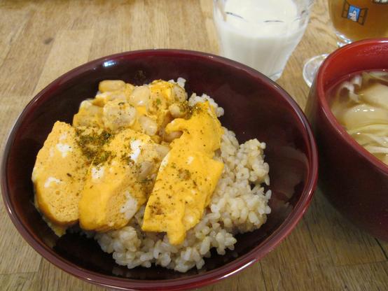 Wooden lacquered bowl containing brown rice topped with sliced Japanese omelettes incorporating soybeans and sprinkled with shichimi mix spices.  By its side another bowl of miso soup with sliced scallions and fu. Behind them a glass of soy milk, and another of dark tea.