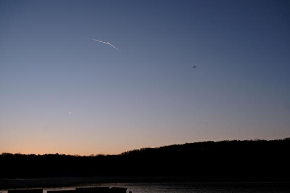 At dawn, a total of five airplanes are in the sky: four contrails, and one visible plane. Sky is blue to light yellow toward the bottom above the woods on a lake